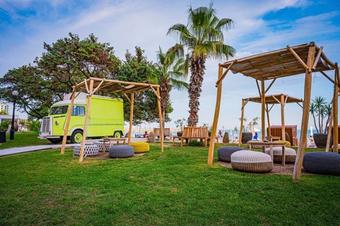 A yellow motorhome next to wooden canopies and palm trees on a green meadow near the sea, Golden