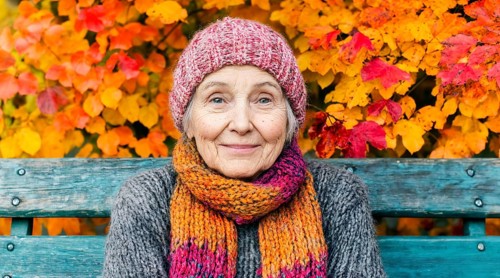 Old woman with colourful knitted hat and scarf sits on a bench in front of autumn-coloured leaves,