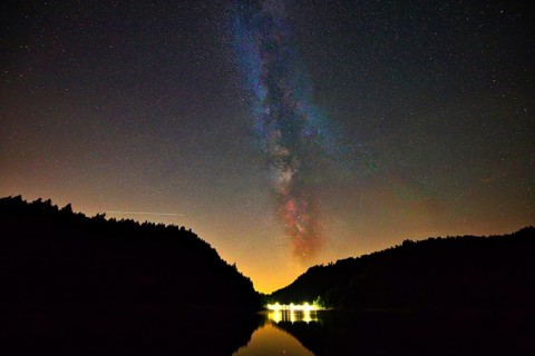 The Milky Way shines in the night sky above a quiet lake surrounded by dark trees, Alp Stausee,