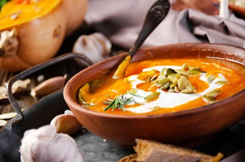 Clay dish with homemade rustic pumpkin soup with seeds on wooden table with bread and greens aside