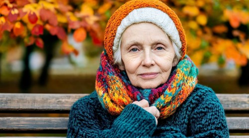 Senior citizen with colourful knitted hat and scarf sits warmly wrapped up on a bench in autumn,