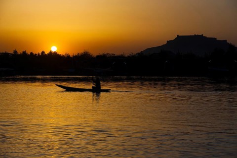 The silhouette of a boat glides along the surface of Dal Lake during sunset with Srinagar Fort in