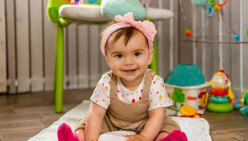 Smiling baby with pink hairband sitting on wooden floor surrounded by toys in children's room, AI