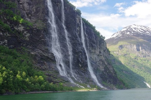 High waterfalls flow down a steep cliff face, surrounded by green forests and mountains under a