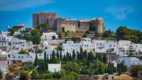 Castle on a wooded hill above white houses and cypresses, under a bright blue sky and warm