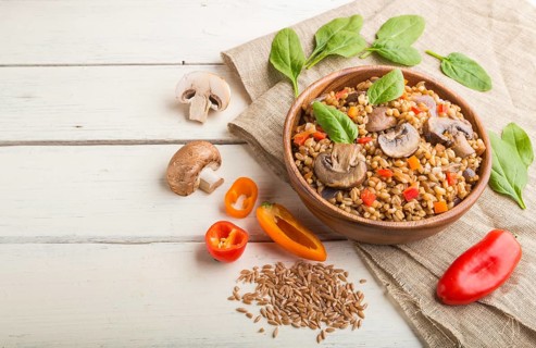 Spelt (dinkel wheat) porridge with vegetables and mushrooms in wooden bowl on a white wooden