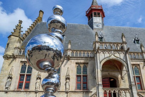 Market square reflected in shiny modern artwork in front of the Brabantine Gothic town hall in the