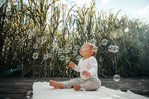 A baby on a blanket surrounded by sunlight, playing with soap bubbles