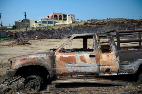 Destroyed lorry in a devastated landscape with burning trees and a building in the background,