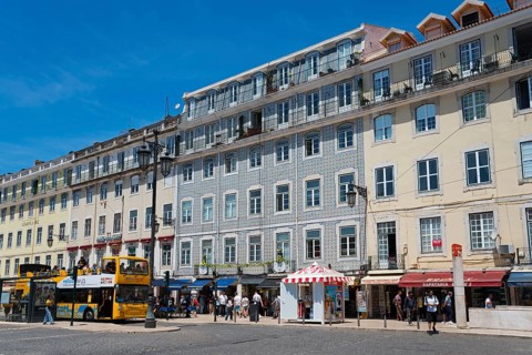 Lively street with many shops and tall buildings, people and a bus under a sunny sky, house with