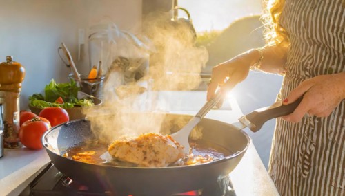 A person is cooking chicken in a pan on a stove. The pan is filled with a red sauce and the chicken