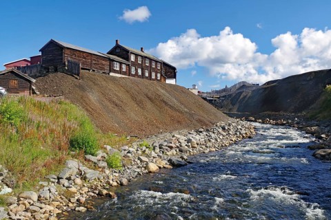 A rushing river next to old buildings on a hill under a sky with white clouds, old mine,
