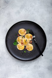Fresh ravioli, with butter, spices and herbs, fried bacon, on a black plate, top view, dark