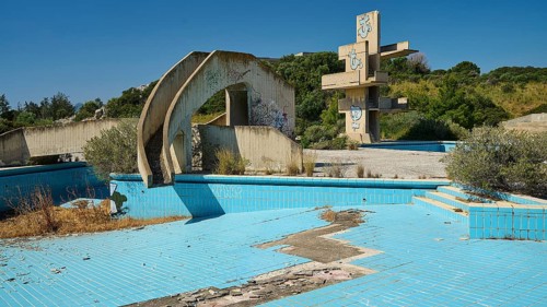 Abandoned swimming pool with concrete ruins and graffiti, surrounded by trees on a sunny day, Lost