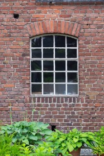 Brick wall with arched window, stable window and lush green plants in front, North