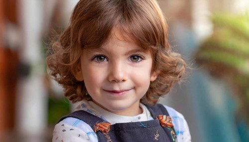 A smiling girl with curly hair wears dungarees and a polka-dot shirt, plants can be seen in the