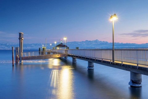 An illuminated jetty over a calm lake with mountains in the background in blue twilight, harbour,