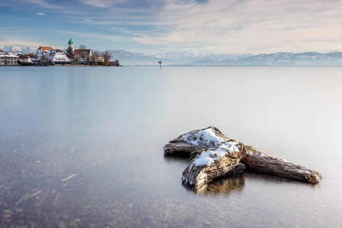 Wooden trunk and picturesque church of St George on the lakeshore in front of snow-covered Pfänder
