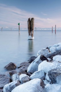 A wooden pile and ice-covered stones on a calm body of water under a blue sky in winter, harbour,