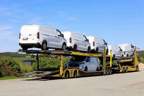 Yellow Volvo vehicle carrier truck transports six new, white Ford Transit Custom vans on trailer,
