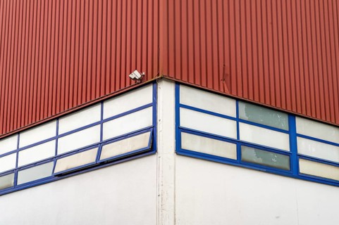 A single surveillance camera is mounted on the corner of a red industrial building with blue-framed