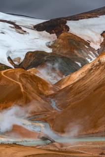 Bridge over steaming stream between colourful rhyolite mountains with snowfields, Hveradalir