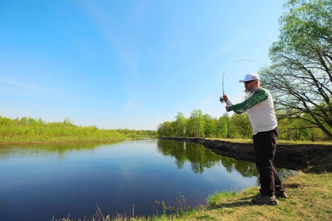 Fisherman with a spinning rod catching fish on a river at sunny summer day with green trees at