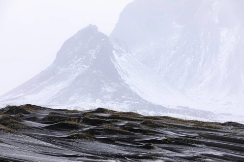 Black sand dunes with reed remains, behind them snowy rocky slopes of Klifantindur, near