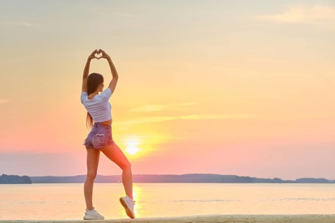 Woman raised her hands up and folded her palms in shape of heart against backdrop of sunset
