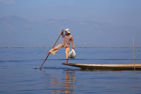 Intha fisherman steering traditional fishing boat by wrapping his leg around the oar, Inle Lake,