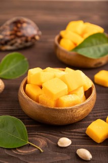 Dried and candied mango cubes in wooden bowls on brown wooden textured background. Side view, close