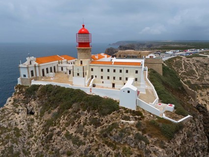 A red lighthouse on a rocky cliff, surrounded by white buildings, with a dramatic view of the