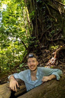 Young man with headlamp, tourist in tropical rainforest, climbing out of the root of a hollow tree,
