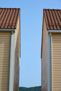 Two wooden houses with red tiled roofs and blue sky in between and mountains in the background,
