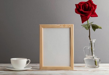 Beige wooden frame mockup with red rose in glass and white coffee cup on gray paper background.