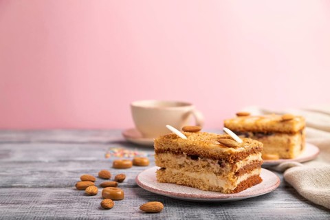 Honey cake with milk cream, caramel, almonds and a cup of coffee on a gray and pink background and