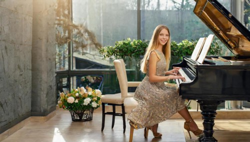 A smiling woman in an evening dress sits at a grand piano next to a bouquet of flowers in the