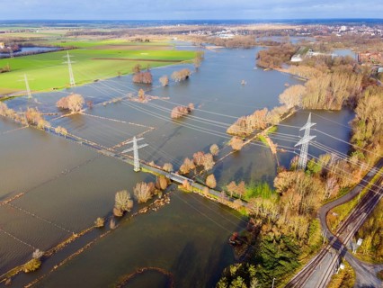 An aerial photograph shows flooded fields and landscape with electricity pylons and cloudy sky,