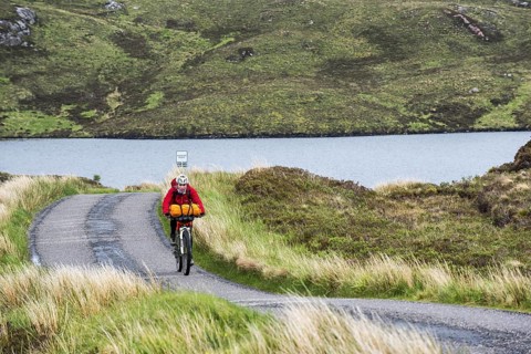 Lonely female biker cycling through the Scottish Highlands on heavily laden touring bicycle along