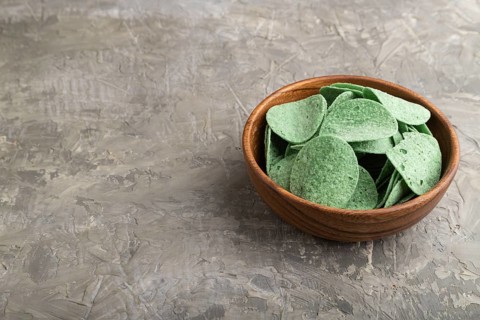 Green potato chips with herbs in wooden bowl on gray concrete background. Side view, copy space