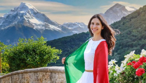 A smiling woman in a white dress holds the Italian flag with a mountain landscape in the