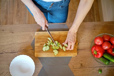 Overhead view of female hands slicing fresh cucumber on wooden chopping board