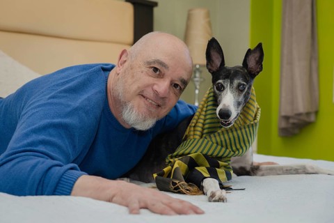 Mature man posing and smiling while looking at the camera with his greyhound pet on the bed