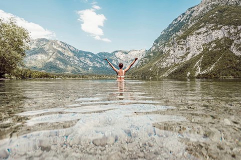 Woman with arms outstretched to the sky on both sides in a clear lake, surrounded by mountains and