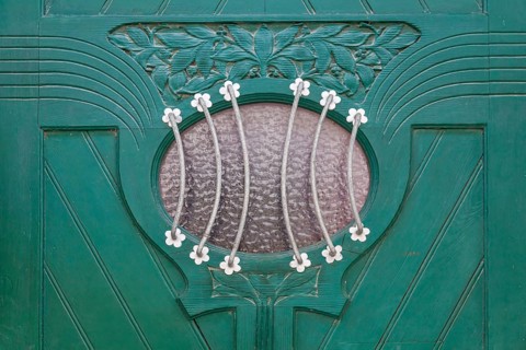 Close-up of a green door with elaborate decorations and a small oval glass window with a metal