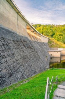 Concrete dam surrounded by green grass and lush forest in the background in sunny weather,