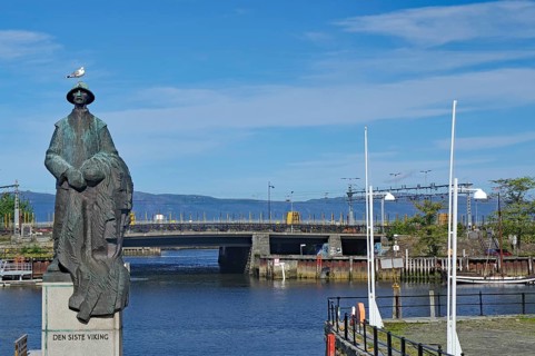 Statue of a fisherman in front of a river and a bridge under a blue sky, the last Viking,