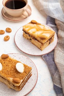 Honey cake with milk cream, caramel, almonds and a cup of coffee on a white concrete background and