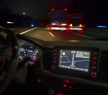 Night-time road scene with two lorries, in front an illuminated cockpit with sat nav of a car,