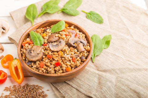 Spelt (dinkel wheat) porridge with vegetables and mushrooms in wooden bowl on a white wooden